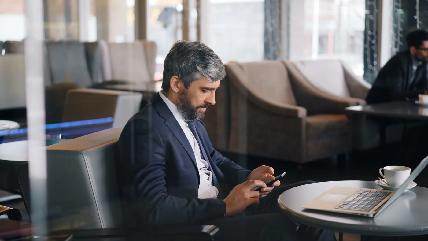 Bir kafede telefonuna bakan takım elbiseli iş insanı; masada dizüstü bilgisayar ve kahve